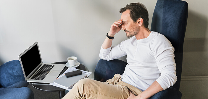 muž v kresle pred počítačom s prázdnou obrazovkou, poznámkovým blokom, šálkou kávy držiacou hlavu za pravú ruku